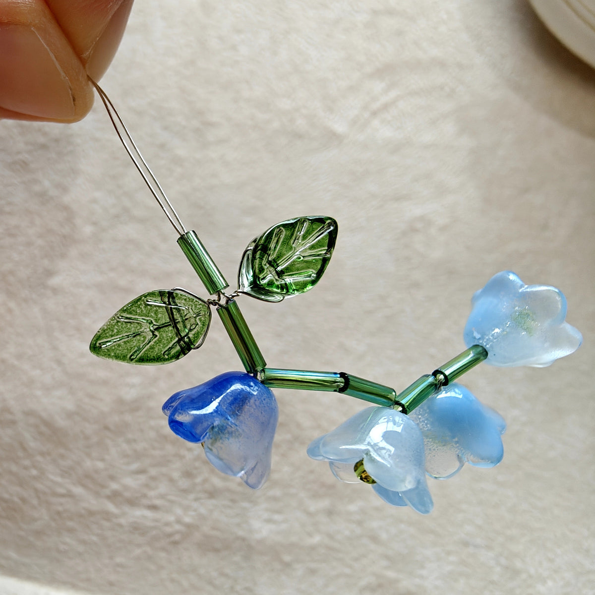 Decorative glass flower with blue petals and green leaves held by a hand on a light background