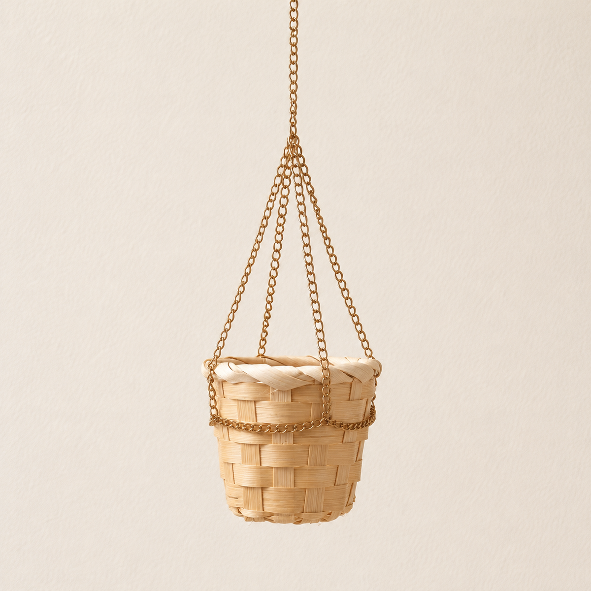 A small woven hanging basket made of light natural wood or rattan, suspended by three gold-toned metal chains against a plain warm off-white wall.