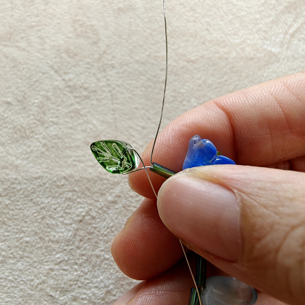 Close-up of a hand holding a needle with a small blue and green bead on a beige background