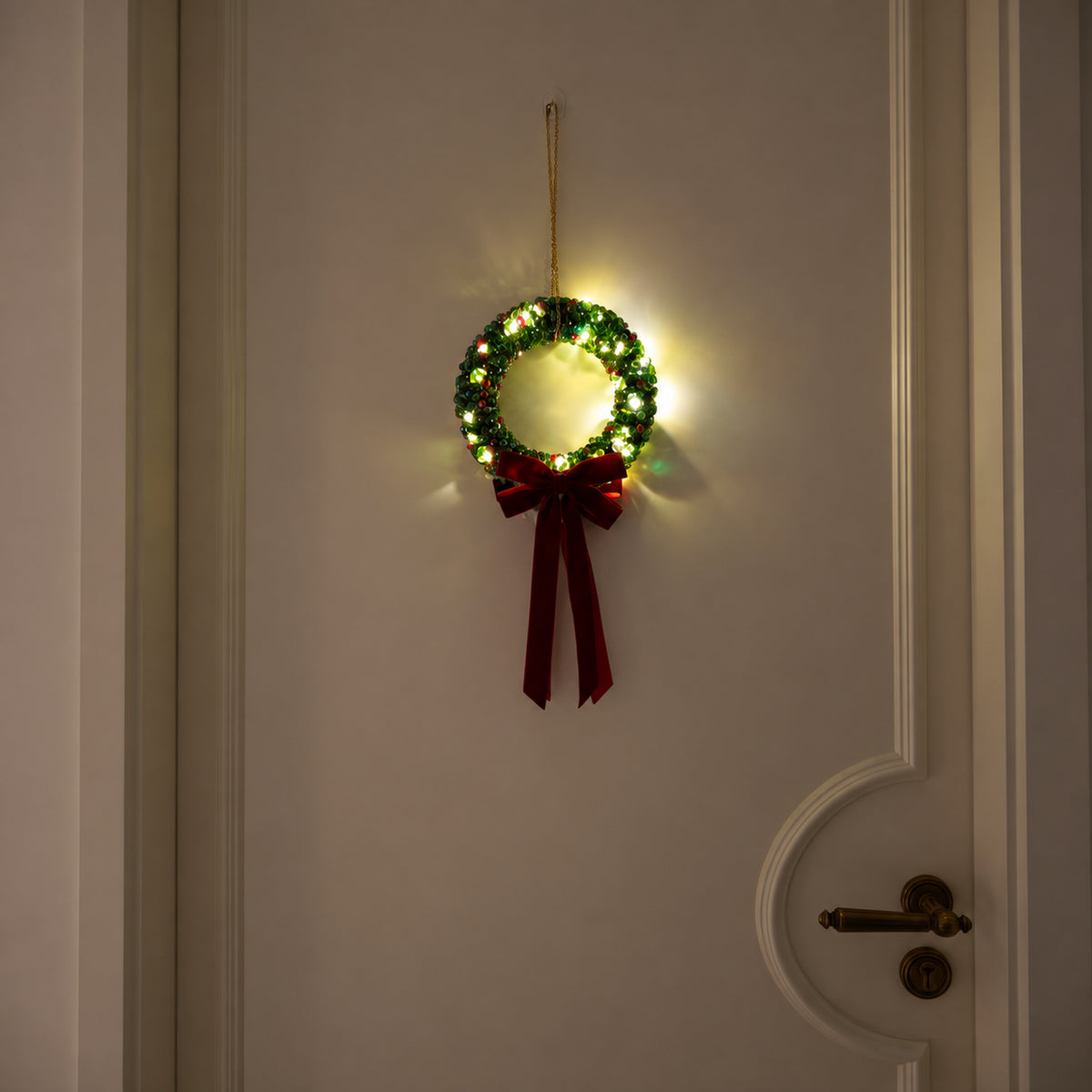 Full green glass-beaded Christmas wreath with warm lights and a burgundy velvet bow hanging on a cream door at night.