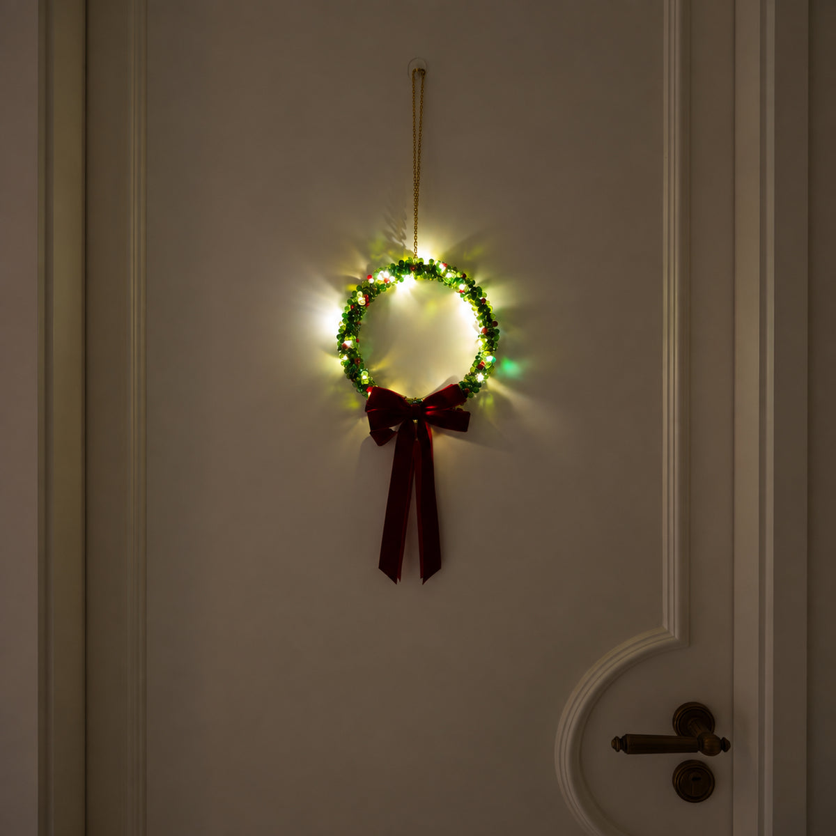 Large slim green glass-beaded Christmas wreath glowing with warm lights and a burgundy velvet bow on a cream door.