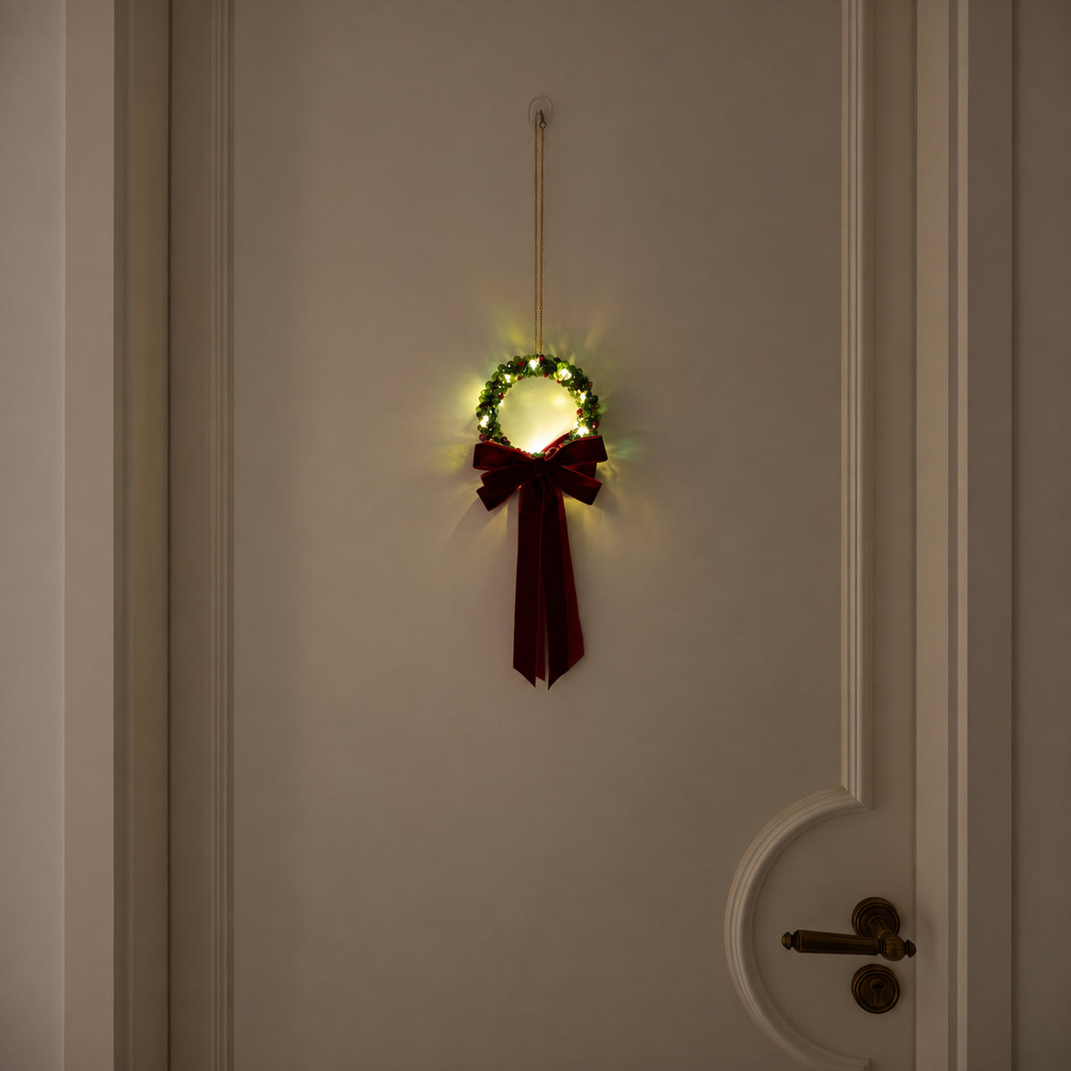 Small green glass-beaded Christmas wreath with warm lights, a gold chain and a burgundy velvet bow on a cream door.