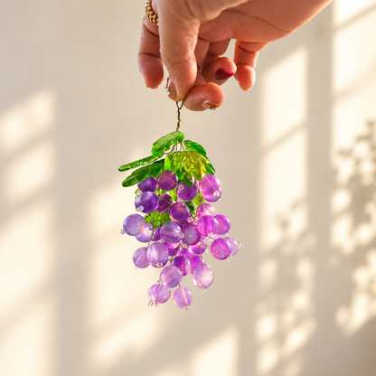 Hand holding a purple beaded grape bunch with green leaf beads against a cream background