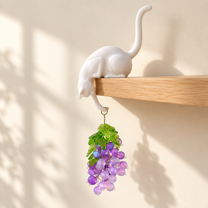 White cat shelf sitter holding a purple beaded grape bunch on a sunlit wooden shelf