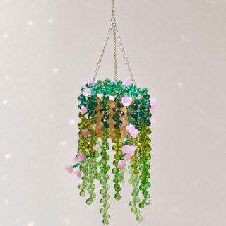 A hanging beaded plant ornament with green and light green faceted beads, pink flower accents, and a gold-toned center, suspended by chains against a light neutral background.