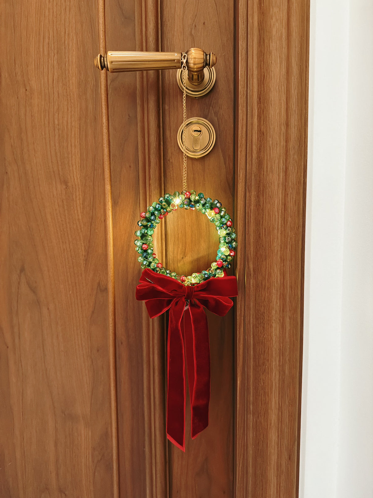 A festive Christmas wreath hanging from a brass doorknob on a wooden door. The wreath is decorated with green and red berries and has a large red velvet bow tied underneath.