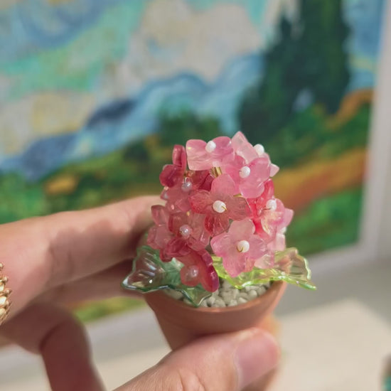 Hand holding a mini beaded hydrangea fridge magnet in a terracotta pot beside a painted landscape
