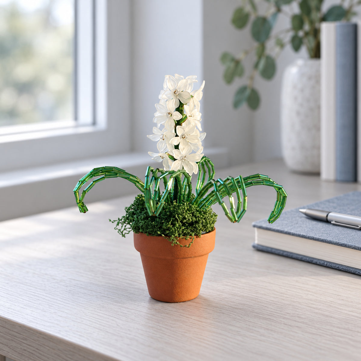 White beaded hyacinth in a small reddish-brown ceramic pot on a cool-toned writing desk.